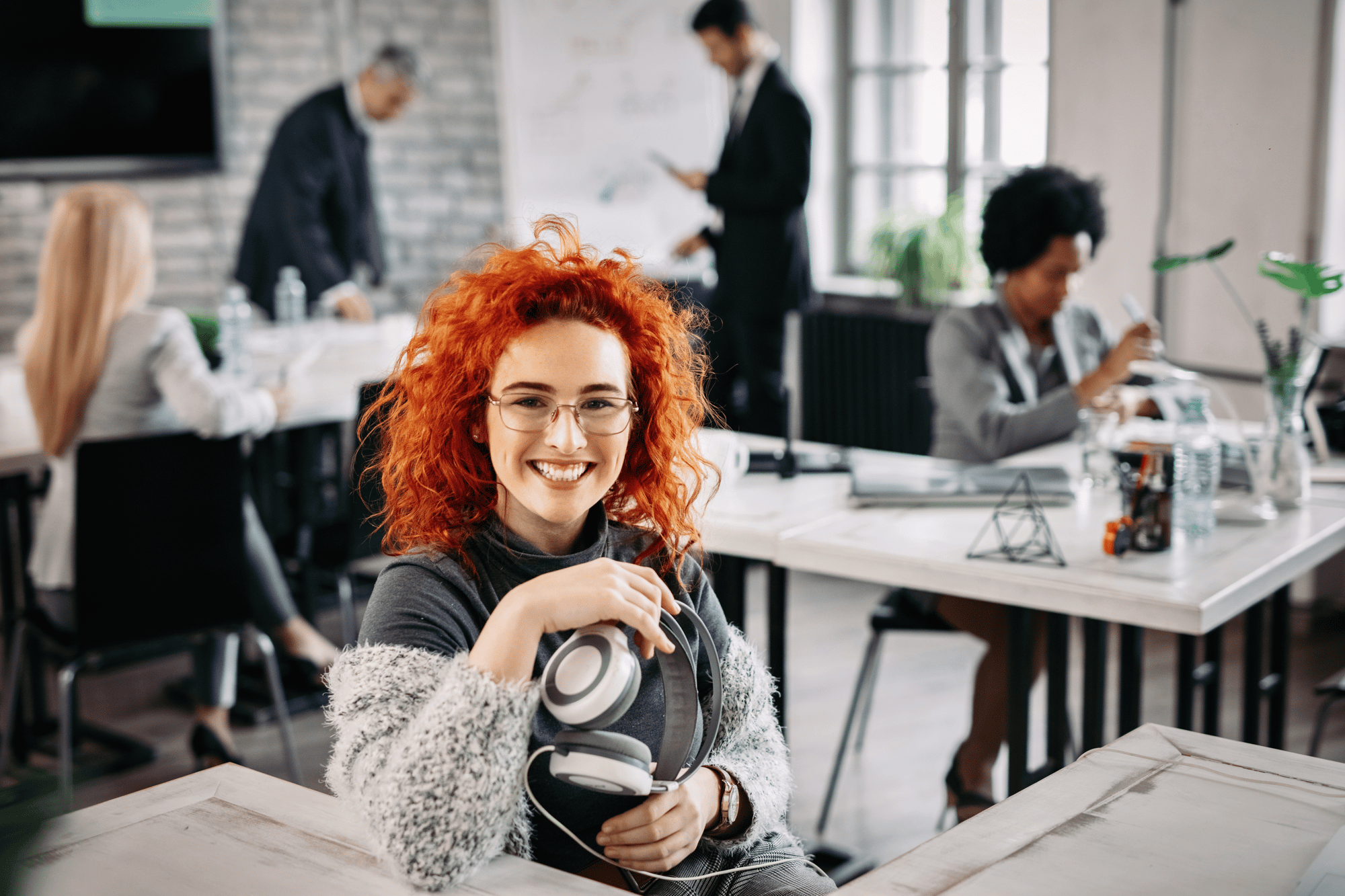 young-happy-redhead-businesswoman-holding-headphones-looking-camera-while-sitting-her-desk-office-her-colleagues-are-working-background-1